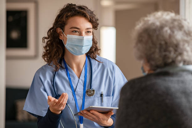 Female doctor holding digital tablet and wearing safety protective mask while talking to senior woman 