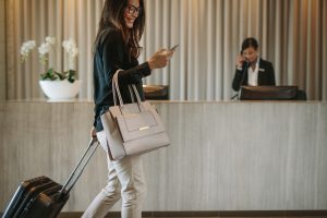 Woman using mobile phone and pulling her suitcase in a hotel lobby. Female business traveler walking in hotel hallway.