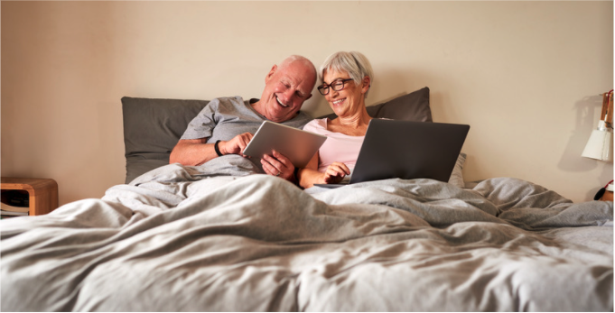 two seniors relaxing in a bed and looking at content on a tablet and a laptop