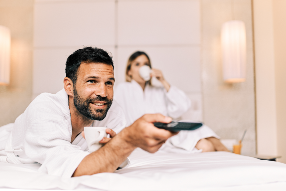 a couple in a hotel room sipping coffee and watching TV